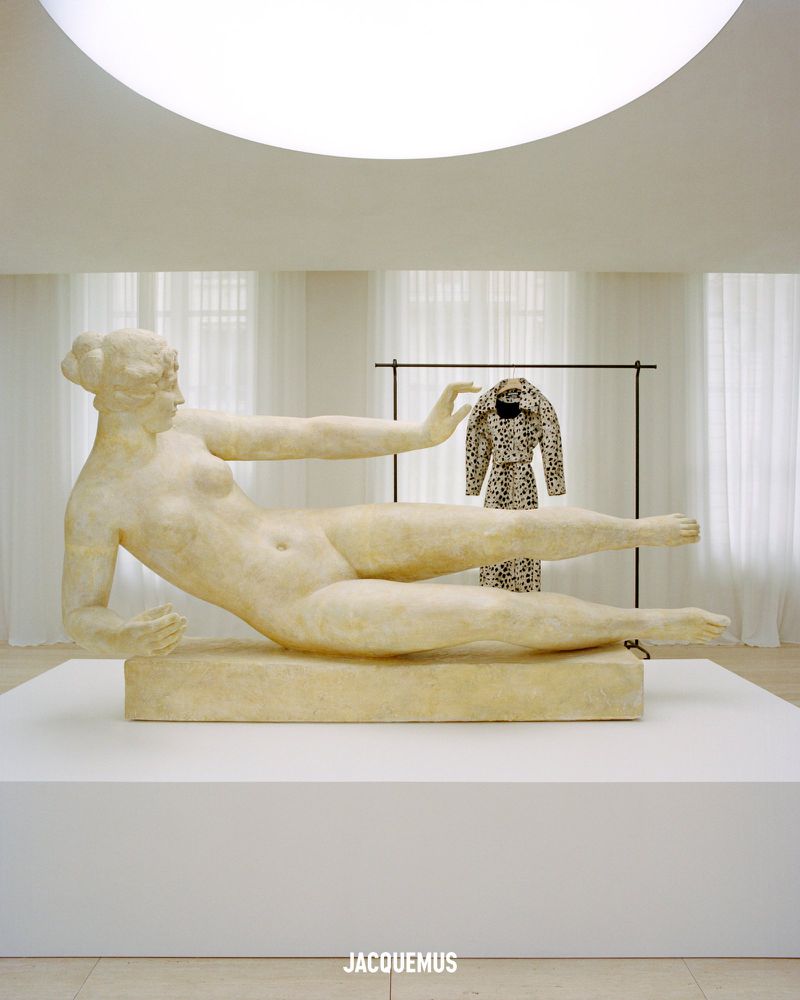 Sculpture of nude female figure laying down on a platform indoors with a dress on a rack in the background.