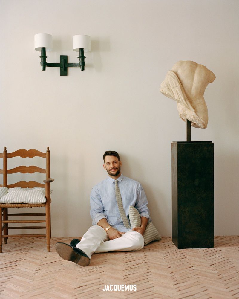 Man in collared shirt and white pants sitting on the floor against a wall, indoors with a marble statue and wooden chair.