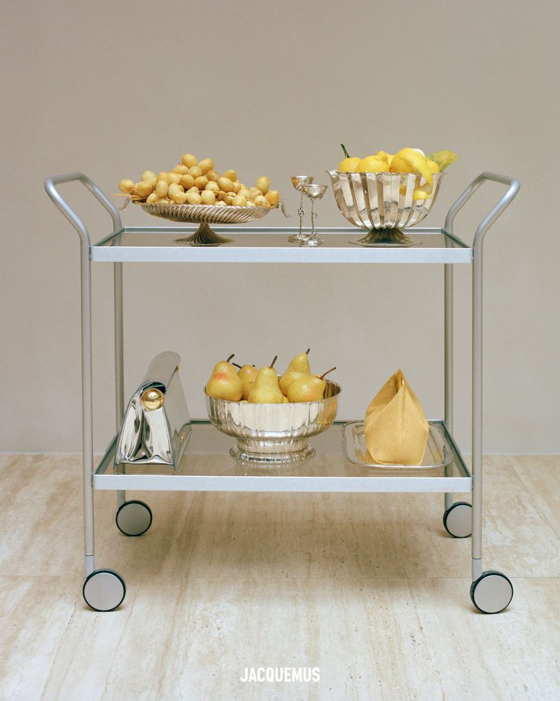 Still life featuring a bar cart with lemons, golden berries, and pears in decorative silver bowls, with a handbag on the bottom shelf.
