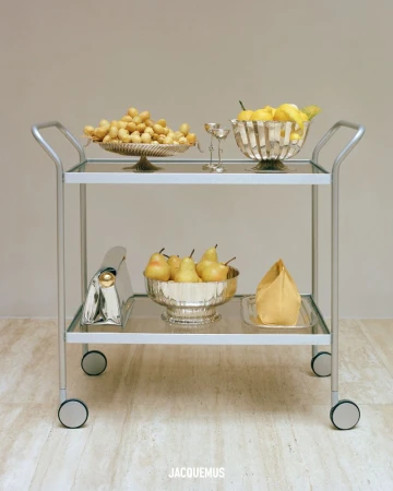 Still life featuring a bar cart with lemons, golden berries, and pears in decorative silver bowls, with a handbag on the bottom shelf.