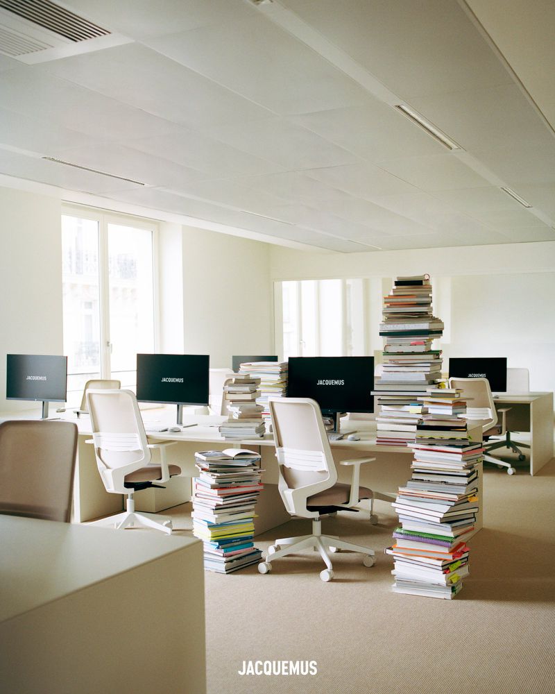 Office space filled with desks, chairs, computers, and stacks of books on the floor. The computers display the JACQUEMUS logo.