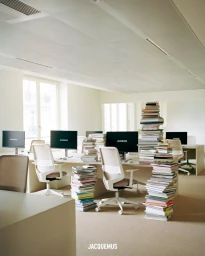 Tall stacks of books standing beside minimalist office desks, white ergonomic chairs, and computer monitors displaying the Jacquemus logo.