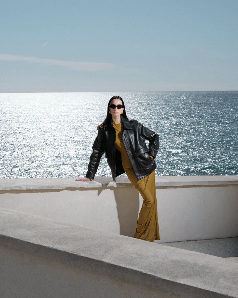 Fashionable woman in leather jacket and long dress poses on a ledge overlooking the ocean on a sunny day. Stylish outdoor portrait.
