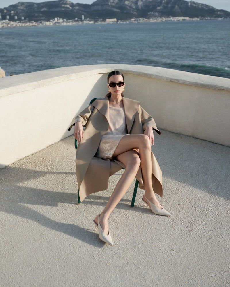 Adult woman sitting in a chair wearing sunglasses and a beige coat with a seascape and mountain background on a bright day.