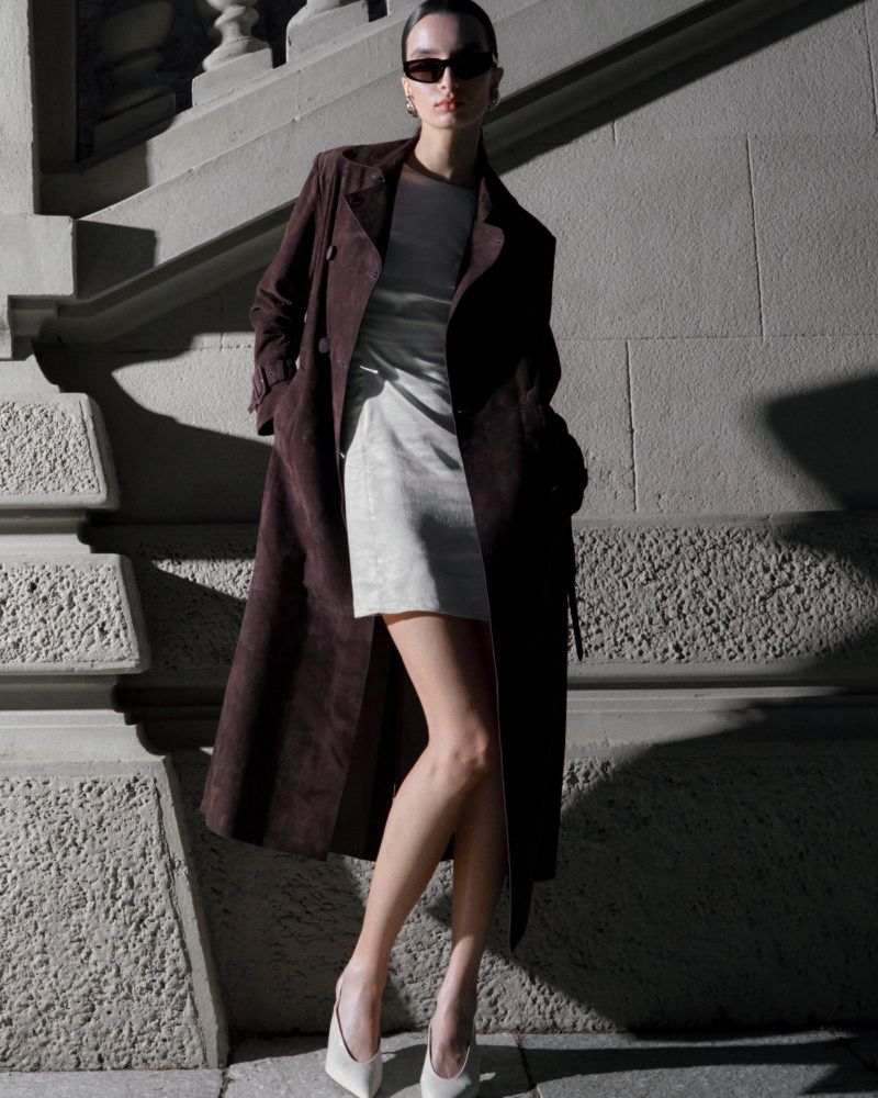 Elegant woman poses on stone steps in brown trench coat, mini dress, and heels, complemented by sunglasses and a stylish gray-toned urban backdrop.