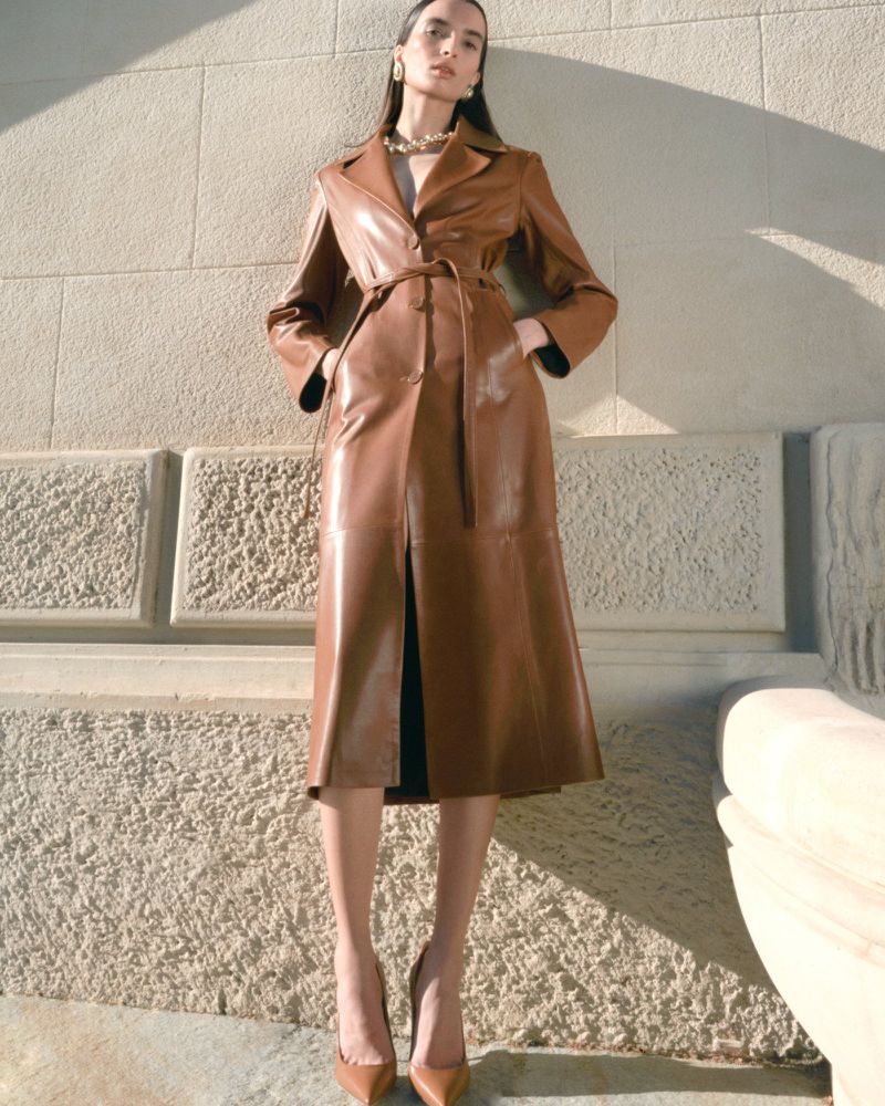 Model Emma Goune wears a belted caramel brown leather trench coat with pointed pumps against a sunlit stone wall for Nour Hammour.