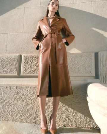 Model Emma Goune wears a belted caramel brown leather trench coat with pointed pumps against a sunlit stone wall for Nour Hammour.