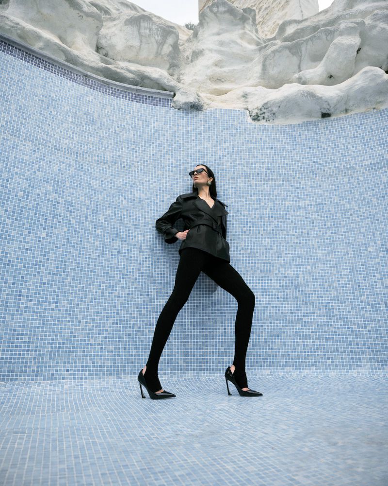 A woman in a black jacket and leggings poses in an empty light-blue tiled pool with a modern architectural backdrop in an urban setting.