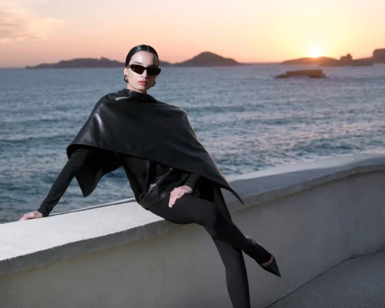 A woman in sunglasses and a cape-style leather jacket poses on a wall with a sea view at sunset, displaying fashion and style.