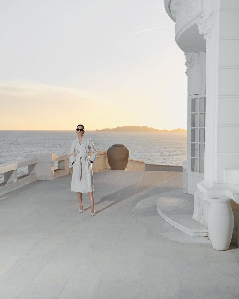 Fashion shot of an adult female model wearing sunglasses and a robe, standing on a balcony overlooking the ocean at sunset.