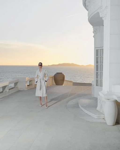 Fashion shot of an adult female model wearing sunglasses and a robe, standing on a balcony overlooking the ocean at sunset.