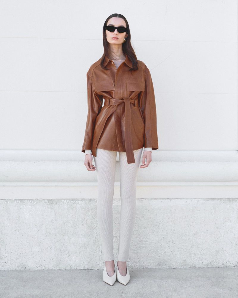 Woman in a brown leather jacket with tie belt, white leggings, and heels, posing against a neutral wall with classic sunglasses.