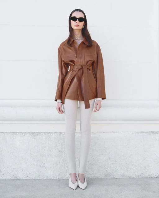 Woman in a brown leather jacket with tie belt, white leggings, and heels, posing against a neutral wall with classic sunglasses.