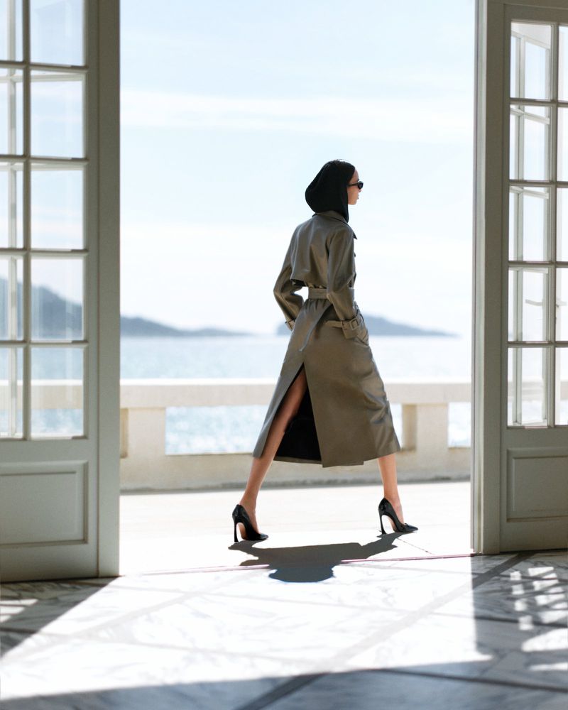 An adult woman walks through an open doorway in a tan trench coat and black heels, toward a sunny view of the ocean on a bright day.