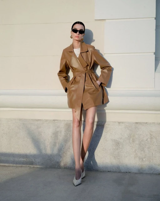 A woman in a tan leather-like trench coat and sunglasses poses against a cream-colored wall in natural light, creating a fashion editorial shot.
