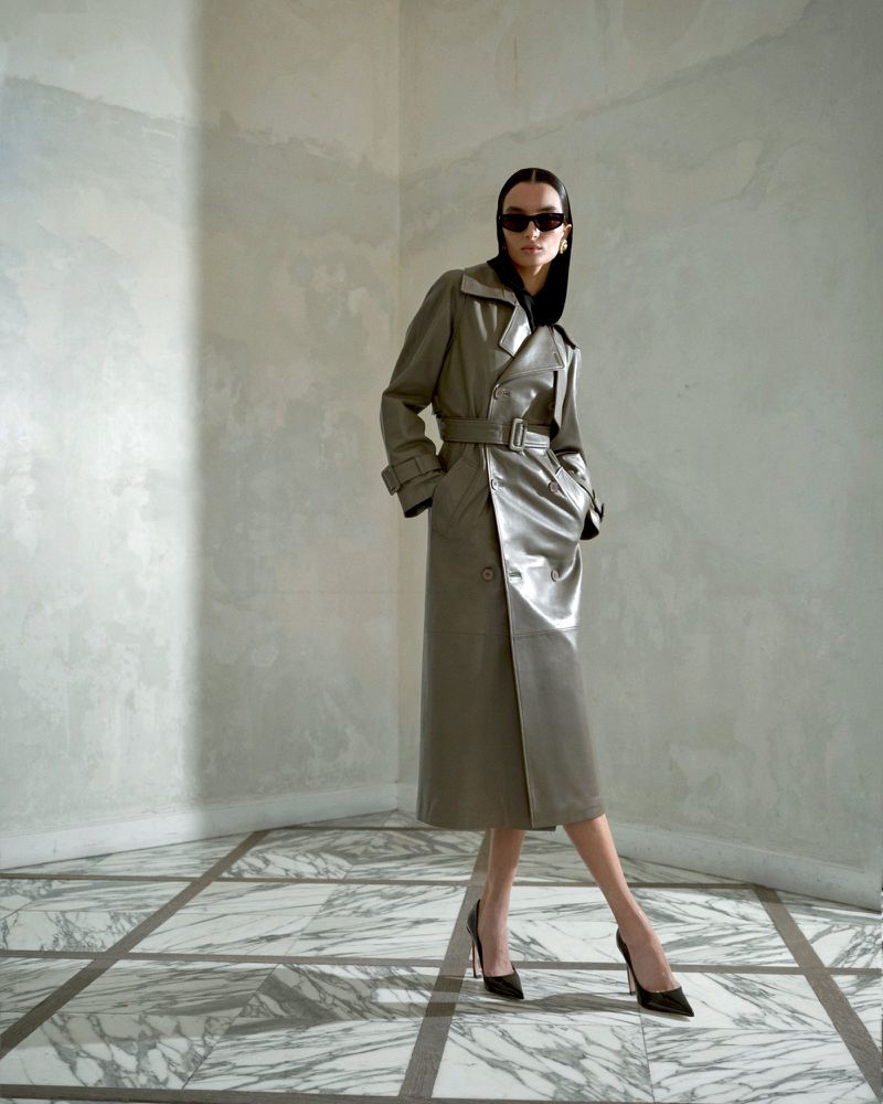 A slender woman models a belted grey trench coat with sunglasses and black high heels in a sparse, tiled interior.
