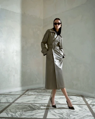 A slender woman models a belted grey trench coat with sunglasses and black high heels in a sparse, tiled interior.