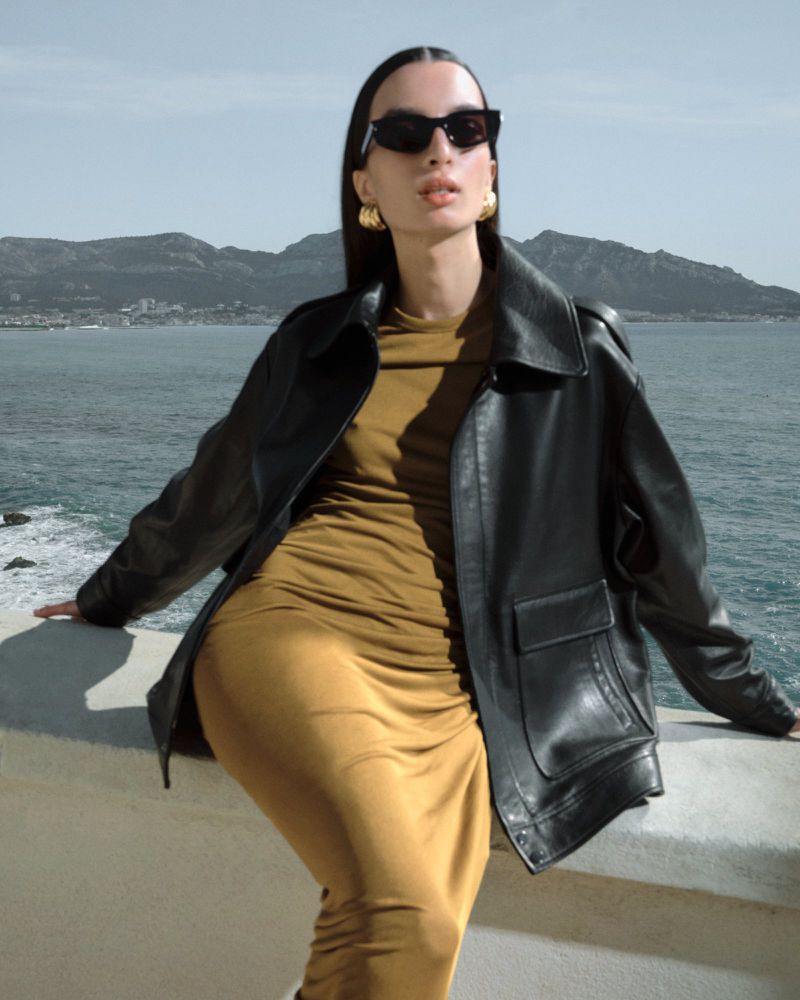 An adult female with black sunglasses and a black leather jacket poses outdoors in front of mountains and blue ocean.