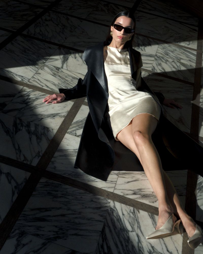 A woman lies on a marble floor wearing a white dress, dark coat, sunglasses, and heels, with strong light and shadow patterns.