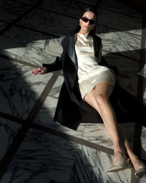 A woman lies on a marble floor wearing a white dress, dark coat, sunglasses, and heels, with strong light and shadow patterns.
