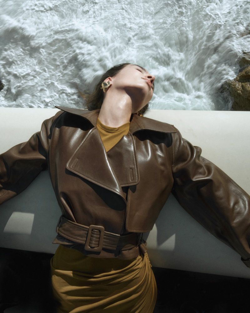 Woman in brown leather jacket lies on white surface with ocean waves visible in the background, illuminated by bright natural light.