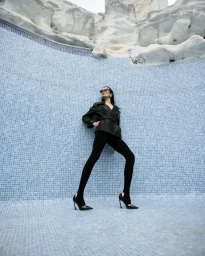 Model Emma Goune posing in an empty blue tiled pool wearing a black leather jacket, stirrup leggings, and stiletto heels.