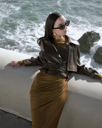 Model leaning against a white ledge by the ocean wearing a brown leather jacket and sunglasses