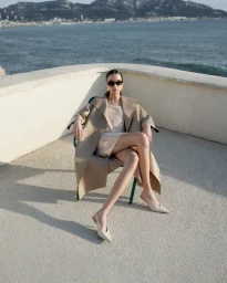 Model seated on a terrace chair wearing a tan coat, shimmery dress, sunglasses, and slingback heels overlooking the ocean.