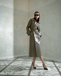 Model in dark sunglasses wearing a long grey belted leather trench coat standing in a room with marble floor tiles.