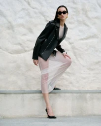 Model wearing a black leather jacket and sheer skirt posing against a textured white wall for Nour Hammour Spring Summer 24.