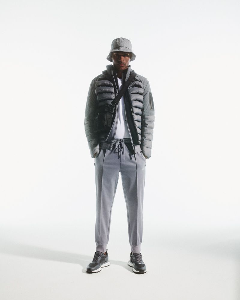 Full body shot of an adult male standing in a photo studio, wearing a grey puffer jacket, bucket hat, joggers, and sneakers.