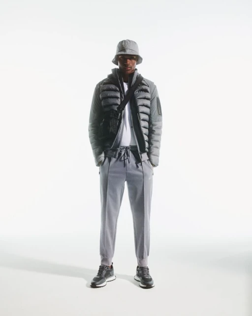 Full body shot of an adult male standing in a photo studio, wearing a grey puffer jacket, bucket hat, joggers, and sneakers.