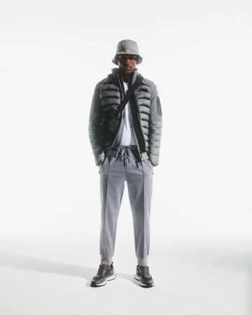 Full body shot of an adult male standing in a photo studio, wearing a grey puffer jacket, bucket hat, joggers, and sneakers.