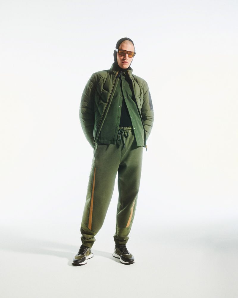 Full-length studio shot of a man wearing an olive green quilted jacket and matching sweatpants with sunglasses and a balaclava.