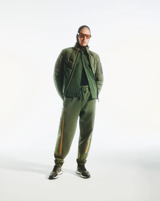 Full-length studio shot of a man wearing an olive green quilted jacket and matching sweatpants with sunglasses and a balaclava.