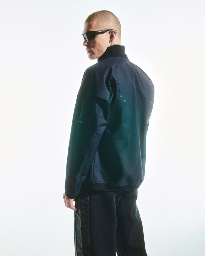 Portrait of a shaved-head adult man wearing a dark jacket and sunglasses, posing in a brightly lit photo studio with a white backdrop.