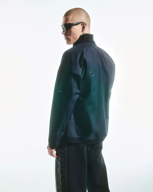 Portrait of a shaved-head adult man wearing a dark jacket and sunglasses, posing in a brightly lit photo studio with a white backdrop.