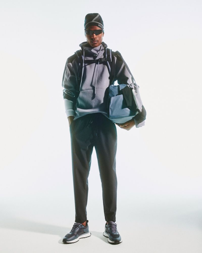 A Black man stands in a gray hoodie, black sweatpants, and sneakers, carrying a blue duffel bag against a bright white backdrop.