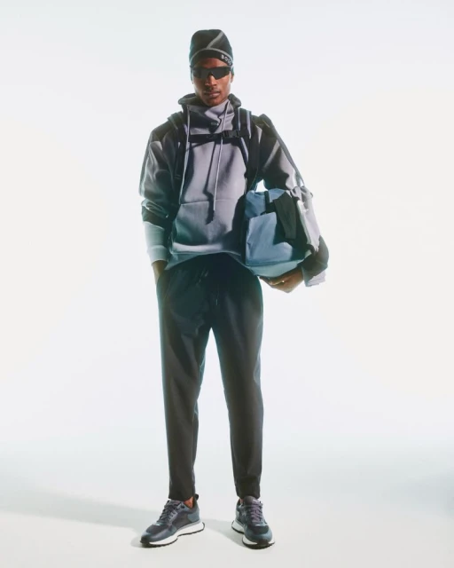 A Black man stands in a gray hoodie, black sweatpants, and sneakers, carrying a blue duffel bag against a bright white backdrop.