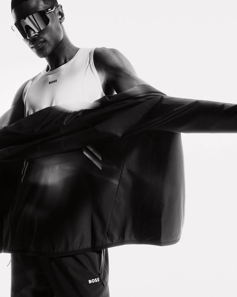 Monochrome studio shot of a Black male model wearing sunglasses, a white tank top, and a black jacket with the BOSS logo.