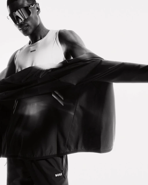 Monochrome studio shot of a Black male model wearing sunglasses, a white tank top, and a black jacket with the BOSS logo.
