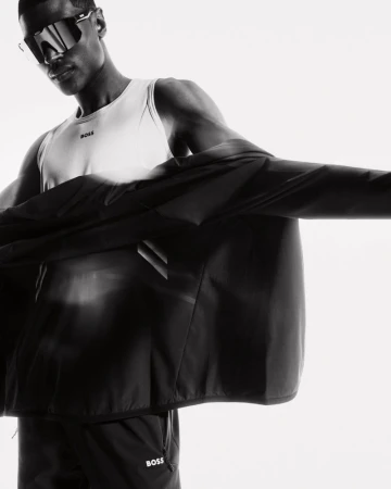 Monochrome studio shot of a Black male model wearing sunglasses, a white tank top, and a black jacket with the BOSS logo.