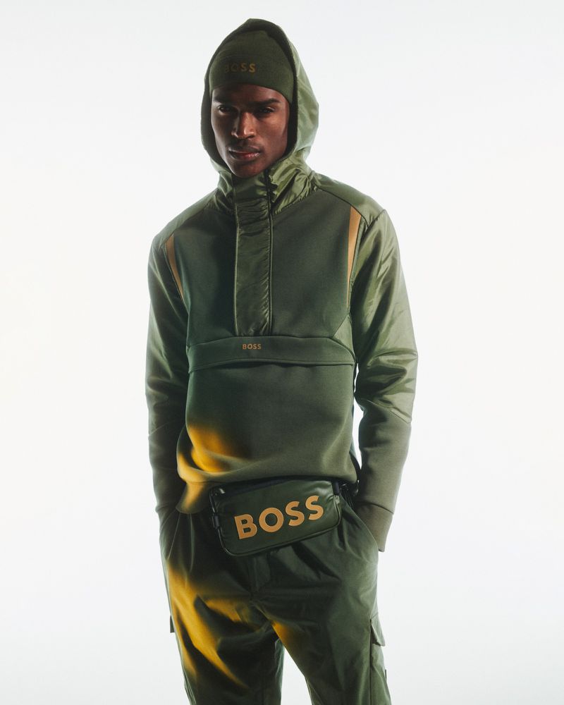 Man in an olive green hooded track suit and matching beanie with the BOSS logo, also wearing a branded fanny pack against a plain backdrop.
