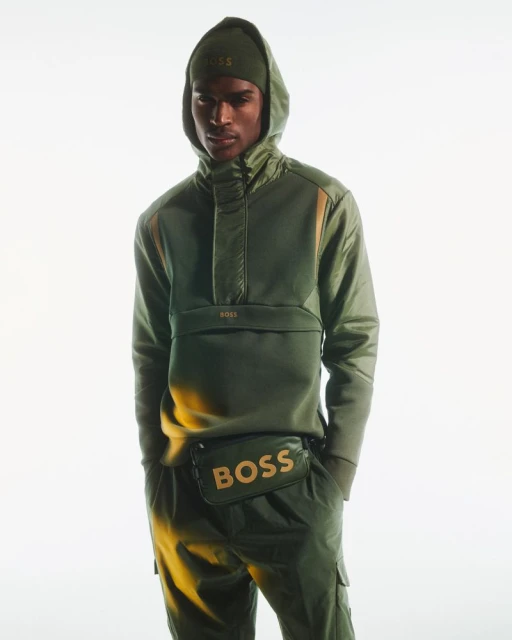 Man in an olive green hooded track suit and matching beanie with the BOSS logo, also wearing a branded fanny pack against a plain backdrop.