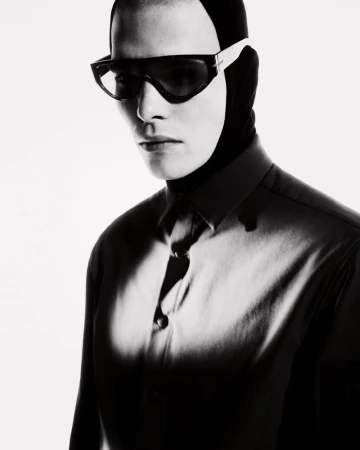 Monochrome portrait of a person wearing futuristic sunglasses and a balaclava-like head covering with a partial view of a collared shirt.