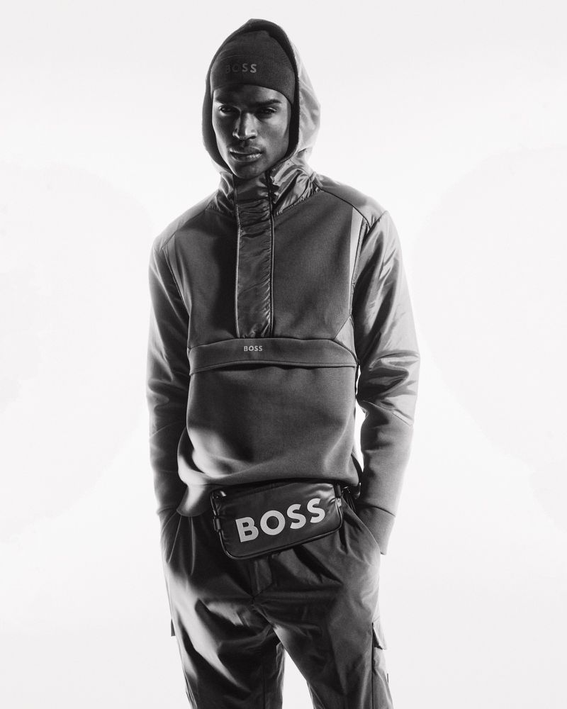 Black and white studio shot of an adult male model wearing a hooded sweatshirt, beanie, and a belt bag with the BOSS logo.