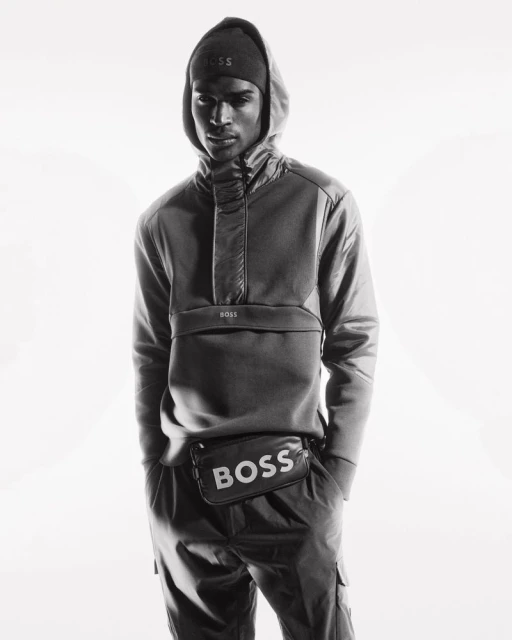 Black and white studio shot of an adult male model wearing a hooded sweatshirt, beanie, and a belt bag with the BOSS logo.