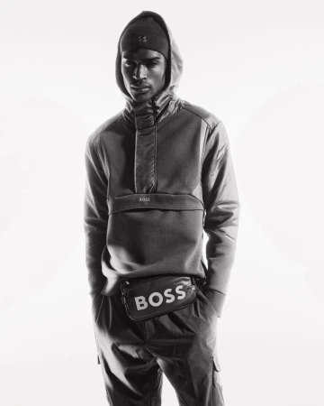 Black and white studio shot of an adult male model wearing a hooded sweatshirt, beanie, and a belt bag with the BOSS logo.