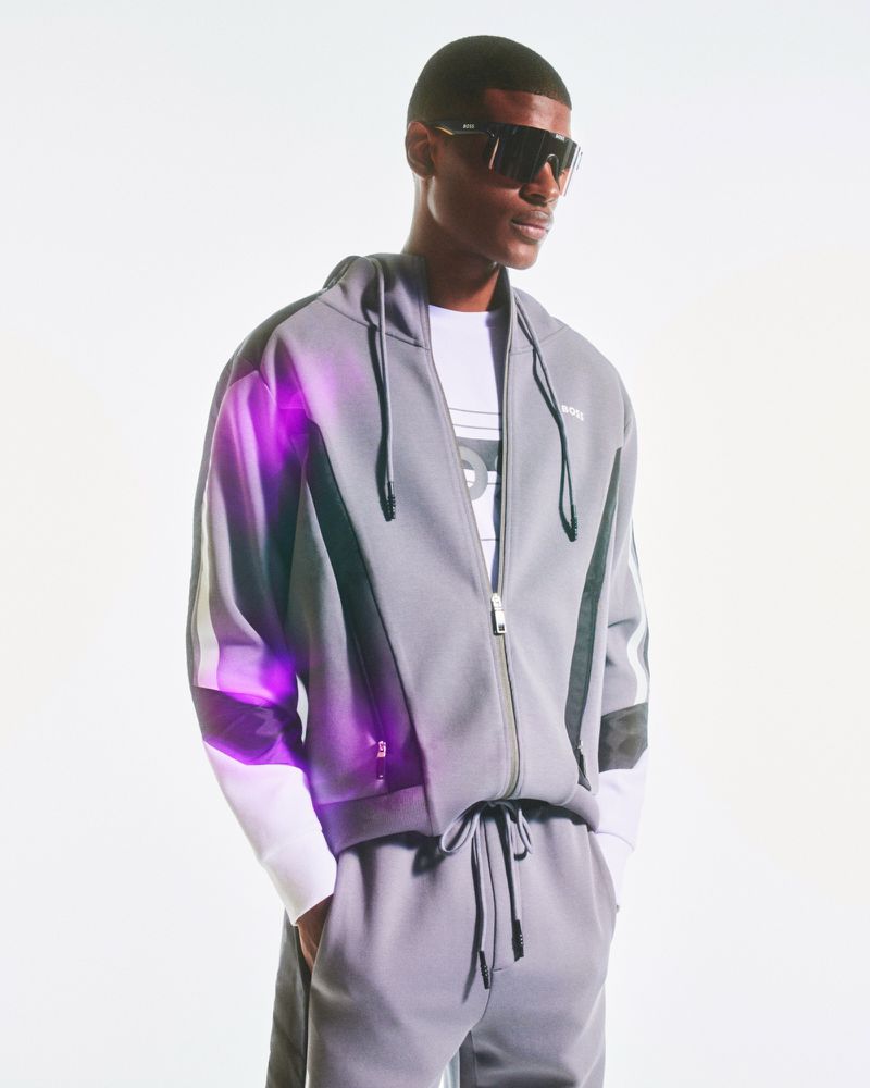 A confident Black male model in a gray tracksuit and sunglasses poses against a bright white background in a studio shot.