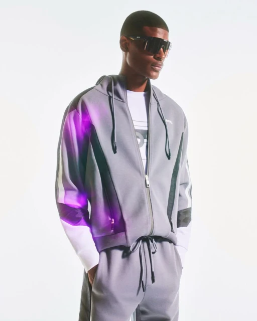 A confident Black male model in a gray tracksuit and sunglasses poses against a bright white background in a studio shot.